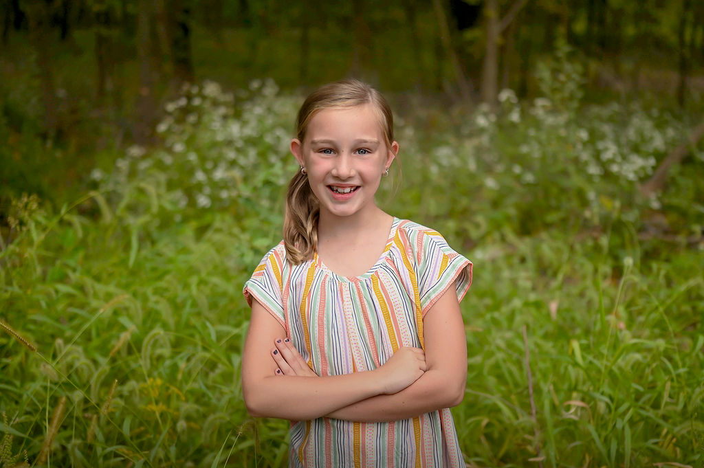 A young girl in colorful stripe dress stands in a forest with arms crossed after visiting Pella Family Dentistry