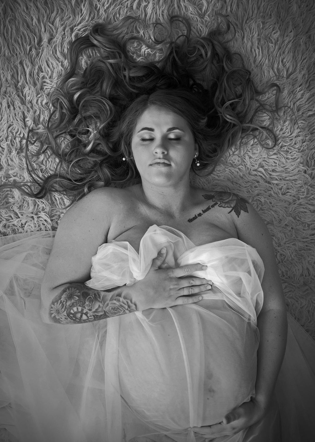 A pregnant woman lays on a shag rug in a studio covering with a sheer white fabric