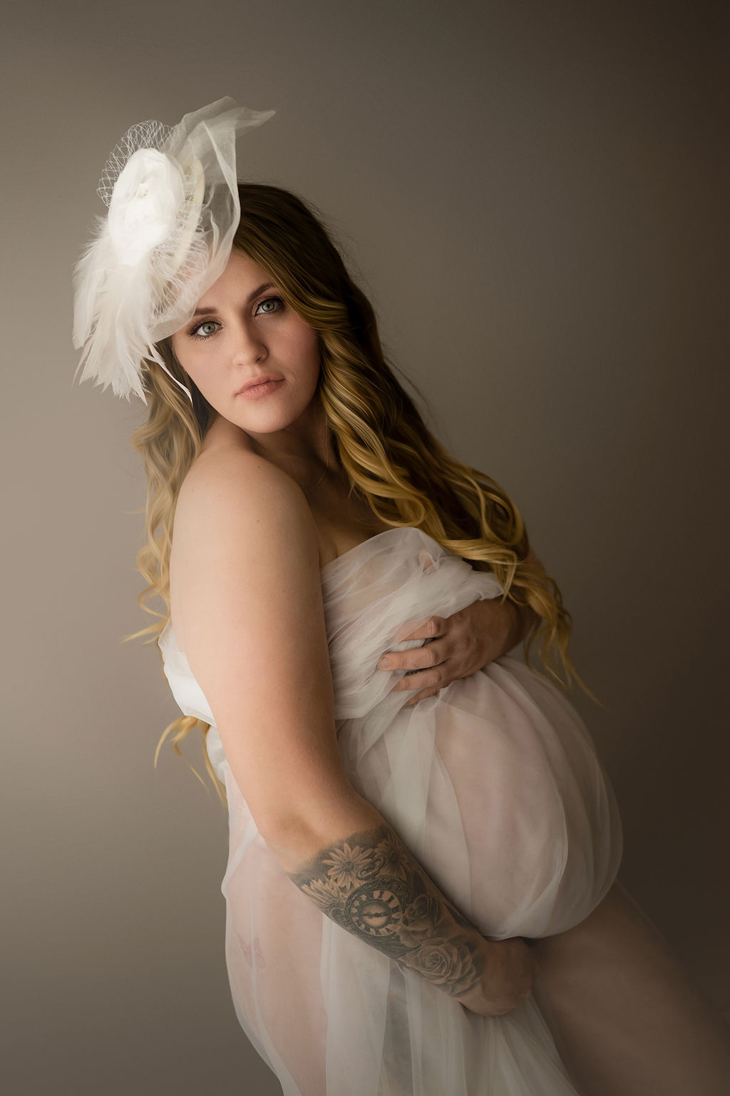 A mom to be covers with a sheer white fabric in a studio with a matching tiny hat after visiting a prenatal chiropractor in des moines