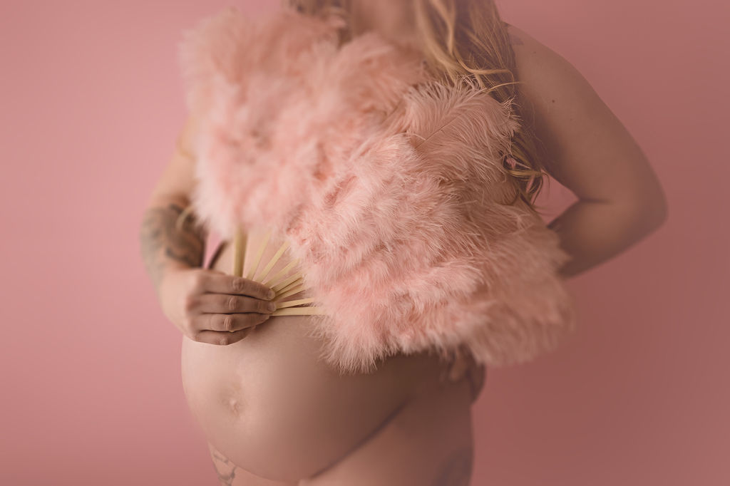 A mom to be stands in a studio covering her chest with a pink feather fan after visiting a prenatal chiropractor in des moines