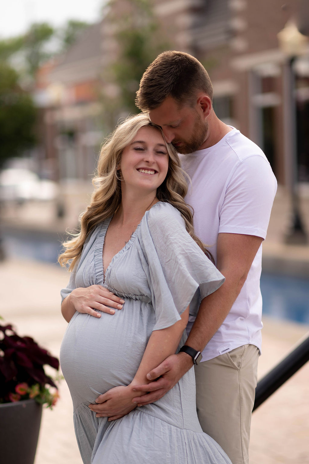 A happy pregnant couple snuggles on a street sidewalk with hands on the bump