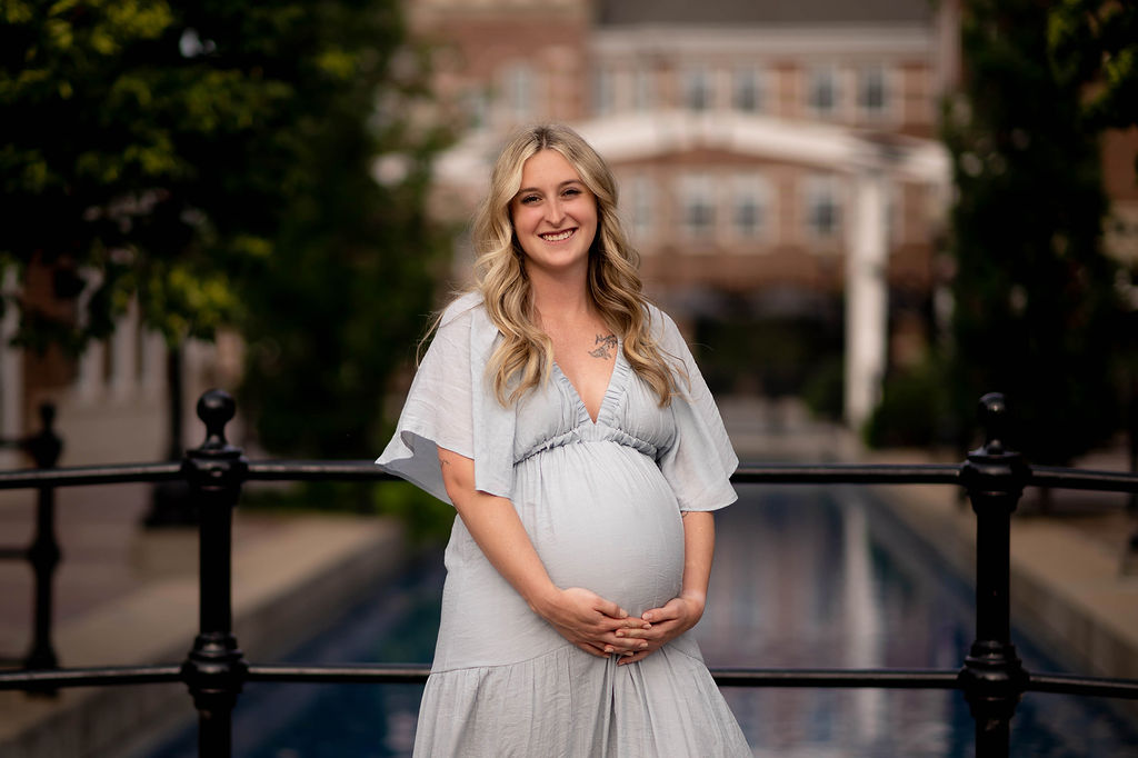 A pregnant woman stands on a bridge over a pond in a blue maternity gown holding her bump after visiting a prenatal chiropractor in Pella, Iowa