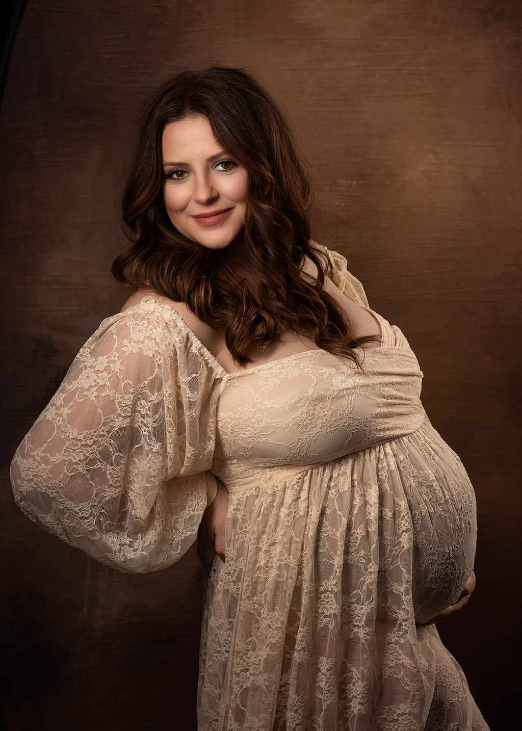 A mother to be smiles while standing in a studio in a cream lace maternity gown after a prenatal massage in Pella, Iowa