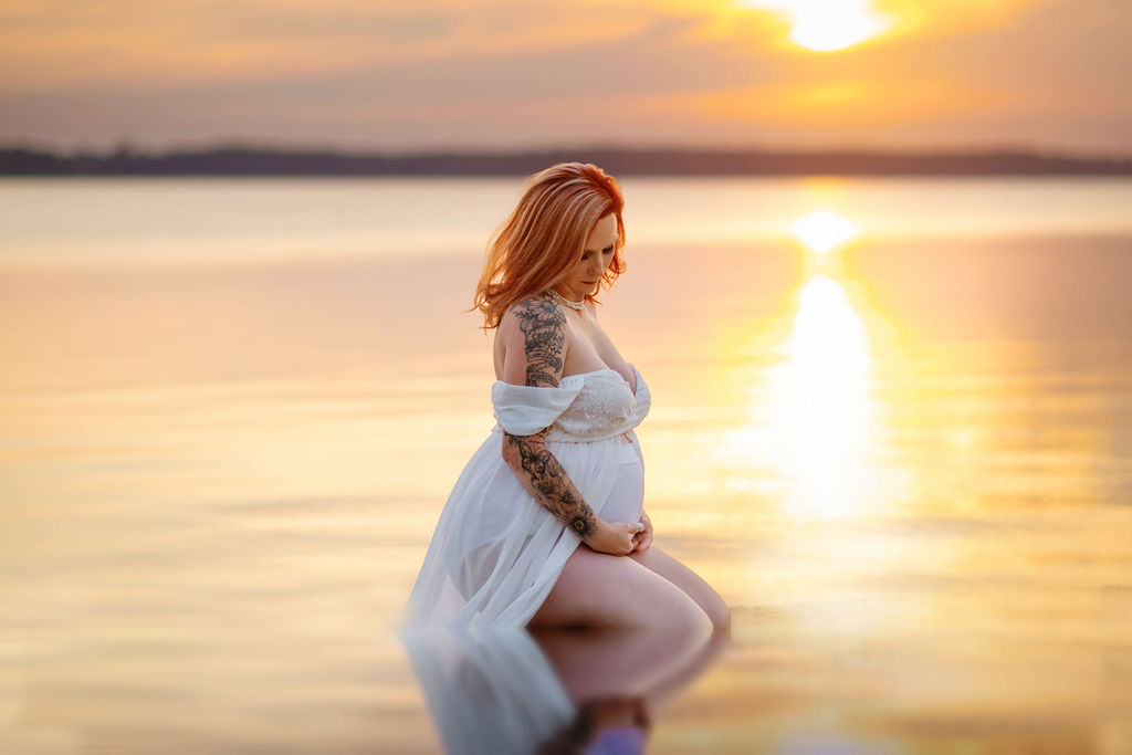 A mom to be kneels in the water at sunset in a white maternity gown