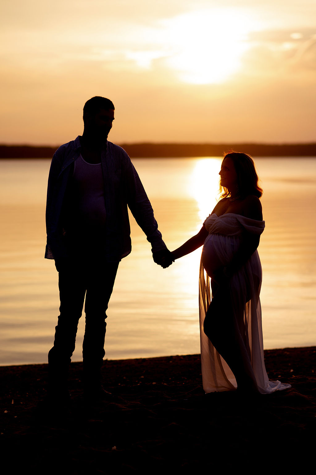 An expecting couple holds hands in silhouette on a beach at sunset after some prenatal yoga in des moines