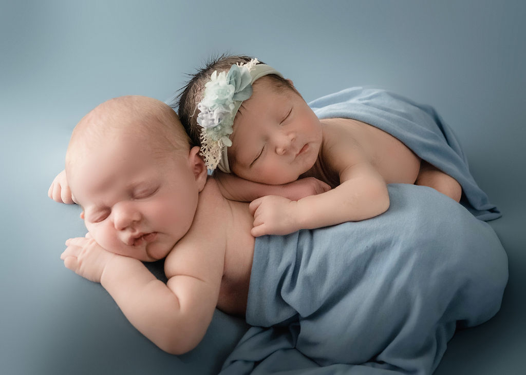 A newborn girl sleeps on top of her twin brother under blue blankets in a studio after visiting purple poppy boutique