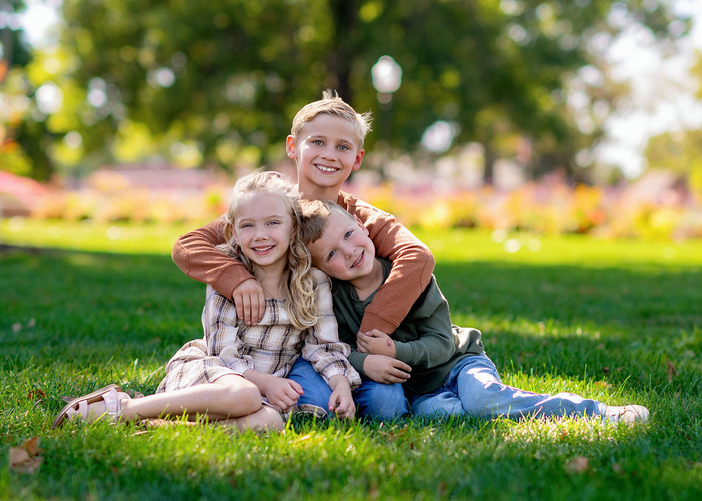 Three young siblings hug and smile while sitting in a park lawn