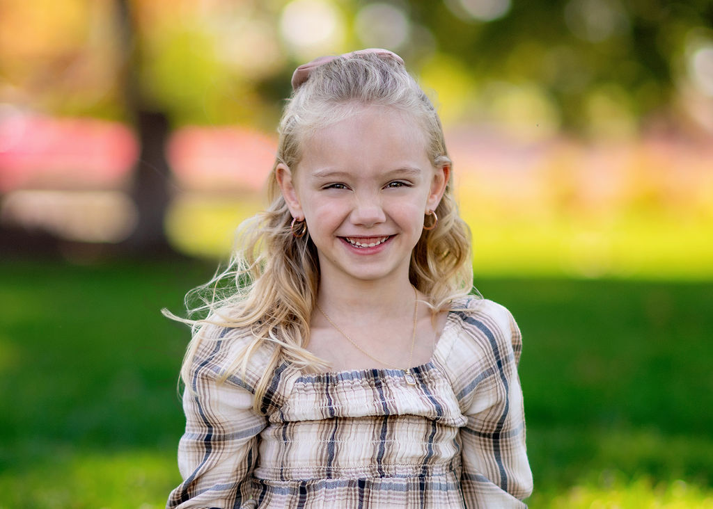 a young girl in a plaid dress stands in a park smiling after visiting Robin Riley